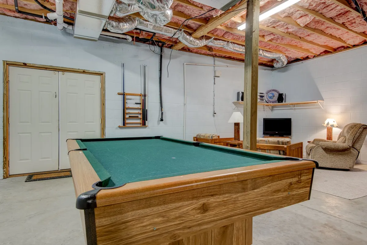 A vibrant downstairs game room scene with a pool table, reclining chair, television, and cozy lighting. The space is set up for recreational play and relaxation, matching the description of the home's entertainment-focused amenities for groups.