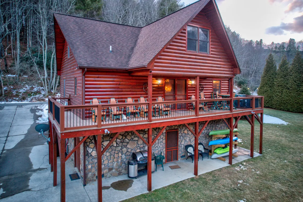 This exterior image features the log cabin structure with an inviting deck furnished for outdoor seating. Prominently visible are kayaks stacked near the deck, a stone foundation, and a dedicated grill area. The scene is set for both outdoor dining and water activities, capturing the adventurous and communal spirit of the porch and patio spaces.