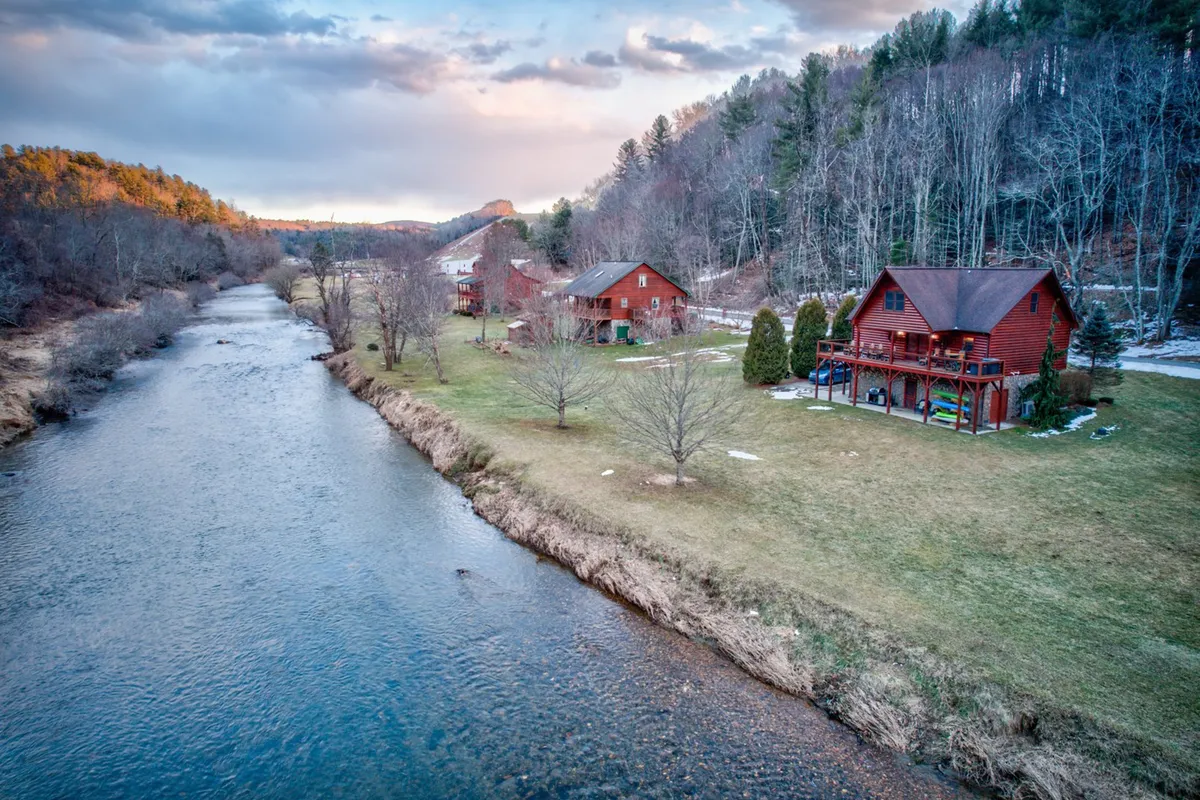 The image showcases a charming red cabin nestled on the banks of the tranquil New River, surrounded by lush trees and framed by rolling North Carolina mountains at sunset. The serene river winds past the property, offering opportunities for fishing and nature walks, while a fire pit near the water invites guests to gather and unwind in the golden evening light.