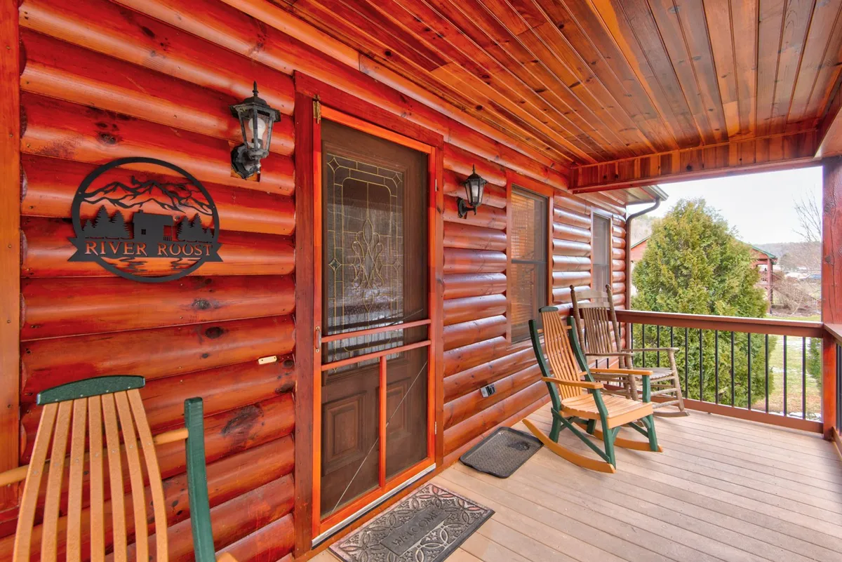 An exterior shot showcasing the front porch of River Roost, complete with wooden log siding, rocking chairs, decorative lanterns, and the welcoming 'River Roost' sign. The scene radiates warmth and the promise of group relaxation.