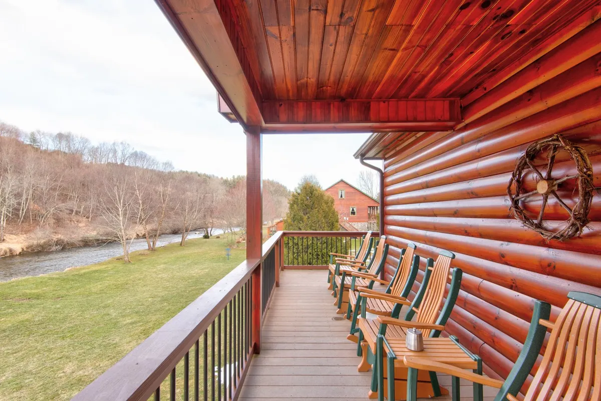This image features wooden rocking chairs on the porch of a log cabin, overlooking a tranquil view of the river with lush greenery and trees surrounding the scene. The calm water is visible, inviting viewers to imagine fishing, kayaking, or tubing right from the riverbank. The overall mood is peaceful and family-friendly, highlighting the direct river access described in the section.