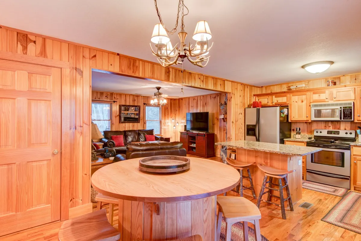 A round wooden table sits between the kitchen and living area, surrounded by warm wood cabinetry and comfortable living room seating. The table is ideal for family meals, board games, or trivia nights, with the kitchen appliances and chandelier visible in the background.