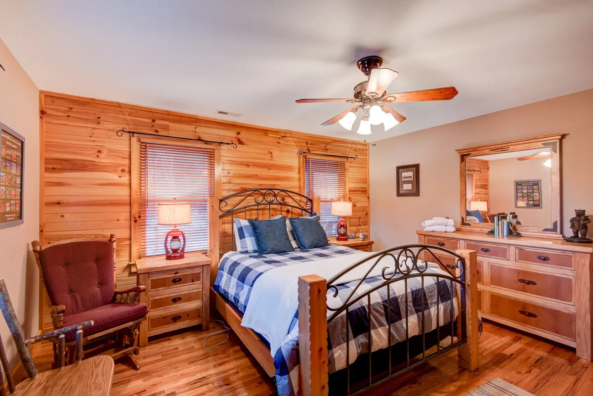 A warm, welcoming bedroom with checkered bedding, a wooden dresser, decorative mirror, and an armchair by the window. The room exudes comfort, offering a tranquil space to unwind and enjoy quiet moments.