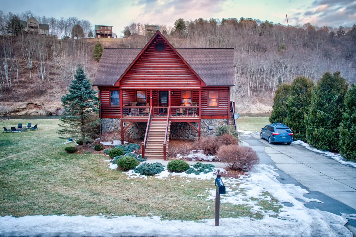 This exterior view highlights the log cabin structure, a welcoming front porch, an evergreen tree, a snow-covered lawn, and the driveway. Activities depicted include relaxing on the porch, gathering around a fire pit, and enjoying outdoor meals, suggesting the perfect post-adventure retreat.