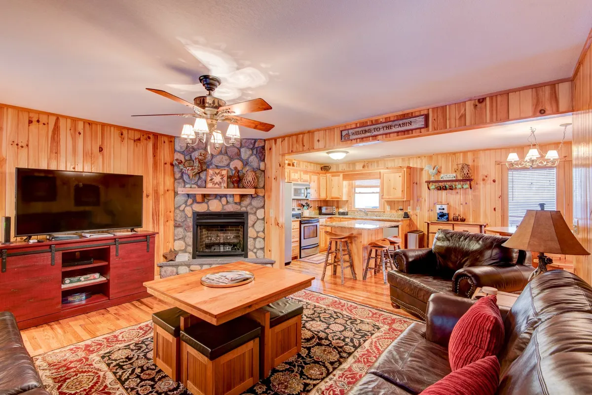 A warm and inviting living room featuring a prominent stone fireplace, a smart TV, wooden furniture, and an adjacent dining area. Plush seating is arranged for comfortable gatherings, and the overall mood is cozy and communal.