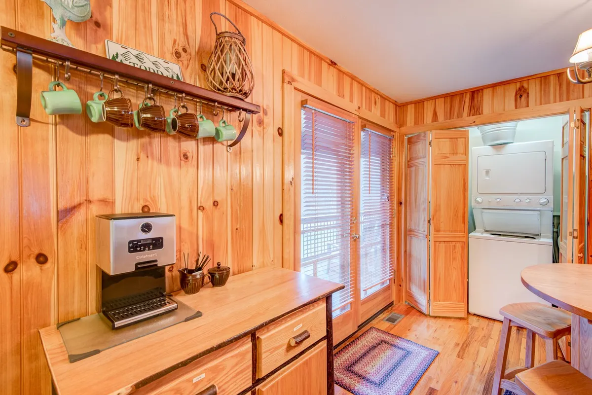 A cozy nook with a coffee machine, hanging mugs, and a wooden dining table set against wooden walls. The inviting setting is perfect for enjoying a morning coffee, reading, or casual conversation.