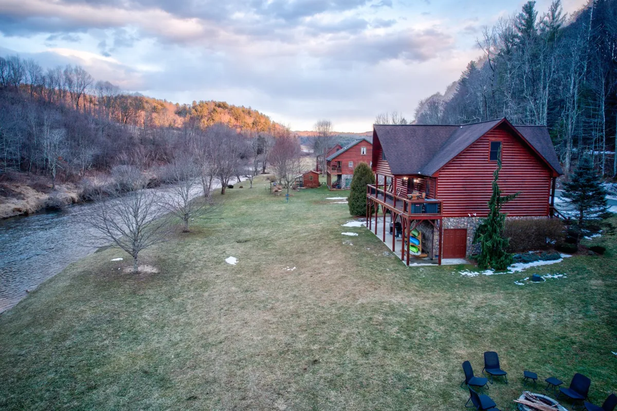 This photo captures a peaceful river scene beside a red log cabin, with trees and an open lawn creating an inviting outdoor space. The riverbank is clearly visible, making it easy to imagine fishing or launching a kayak directly from the property.