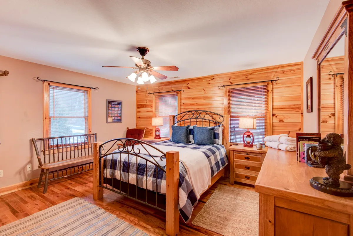 A bright bedroom featuring a comfortable bed with striped bedding, warm wooden furniture, and large windows with blinds that allow sunlight to pour in. The space feels restful and inviting—an ideal retreat for relaxation or a peaceful night’s sleep.