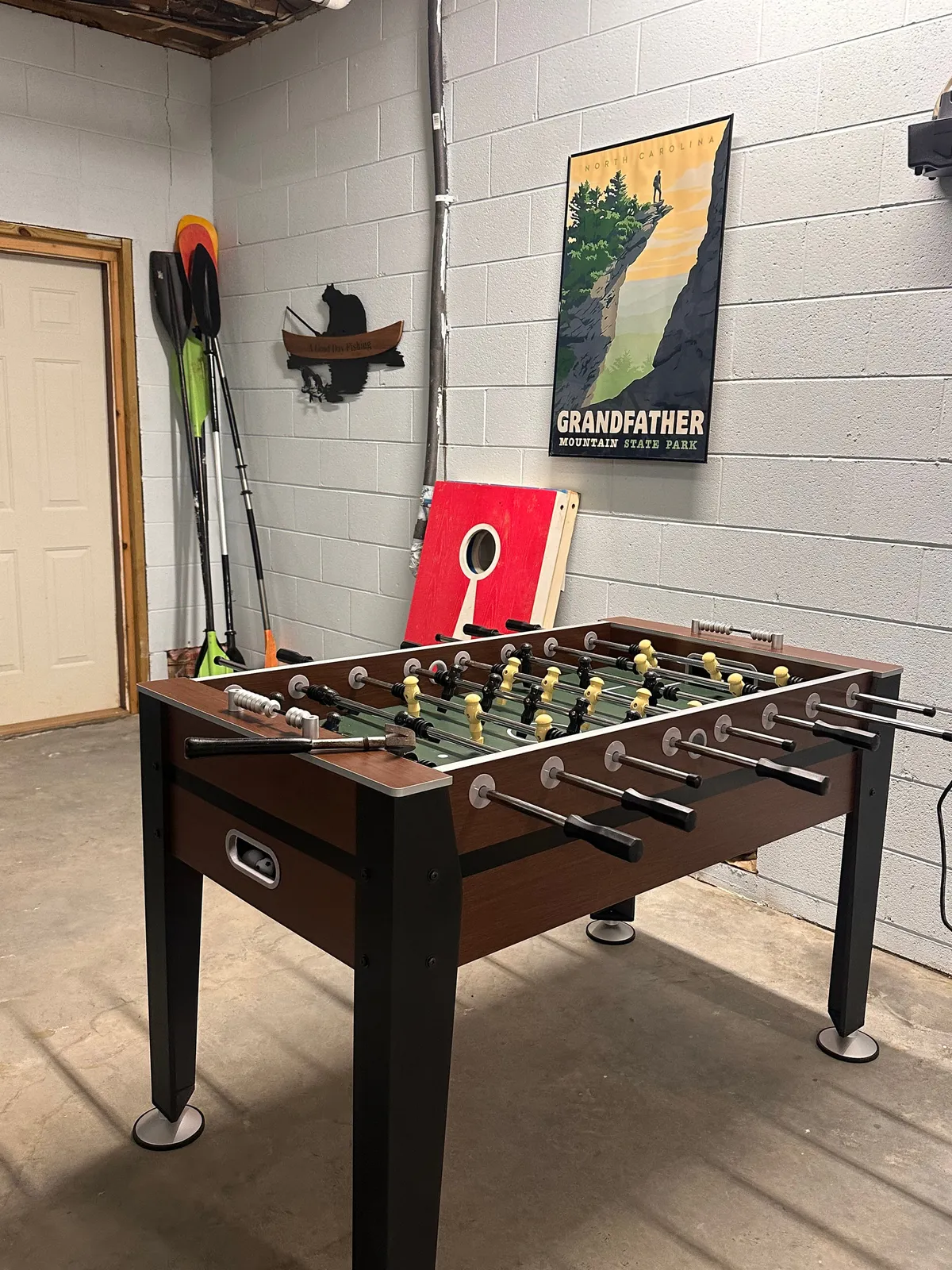 A playful corner of the game room featuring an intense foosball match in progress. The Grandfather Mountain poster and kayak paddles on the wall add unique personality and a touch of local adventure. Friends are gathered around the foosball table, actively engaged and enjoying themselves.
