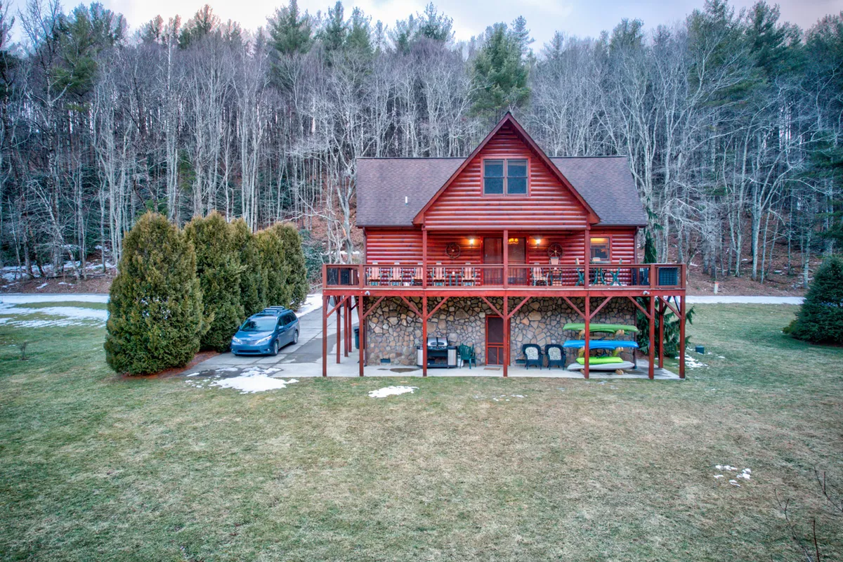 This exterior image shows a log cabin with a large deck, green kayaks ready for use, a front yard with patches of snow, and surrounding trees. The visible kayaks and deck immediately evoke the sense of direct river access and outdoor adventure mentioned in the section narrative.