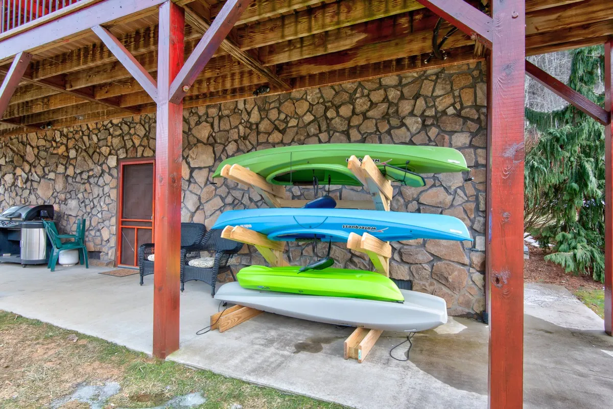 Stacks of colorful kayaks are ready for adventure near a cozy stone wall and patio furniture, set in a lush outdoor area. The scene suggests easy, immediate access to kayaking as a family activity, with comfortable spaces to relax and socialize after an active day on the river.