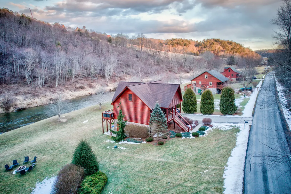 A red wooden house sits beside the gently flowing river, surrounded by green lawns and a comfortable seating area for gatherings. The scene is peaceful yet lively, suggesting both active pursuits (like fishing and riverside picnics) and evening campfire stories or stargazing under open skies.