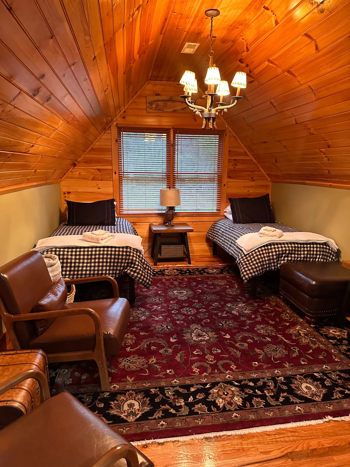 An inviting loft-style bedroom featuring two twin beds with checkered bedding, a wooden chandelier overhead, a bedside table with a warm lamp, and an ornate rug. The space is playful yet cozy—ideal for children to relax or share bedtime stories.